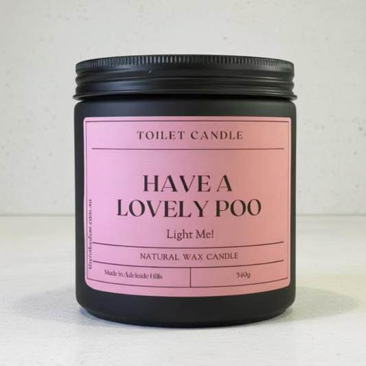 A black glass  jar candle with a pink label reading 'TOILET CANDLE HAVE A LOVELY POO Light Me! NATURAL WAX CANDLE' on a light wood surface against a neutral beige background.
