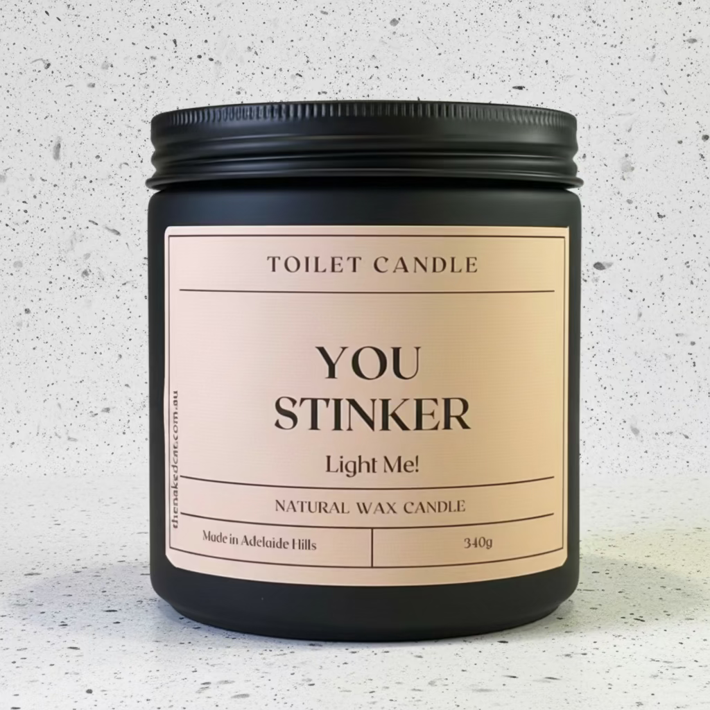 A black glass jar candle with a black metal screw-on lid and a pale pink label reading 'TOILET CANDLE YOU STINKER Light Me! NATURAL WAX CANDLE'.