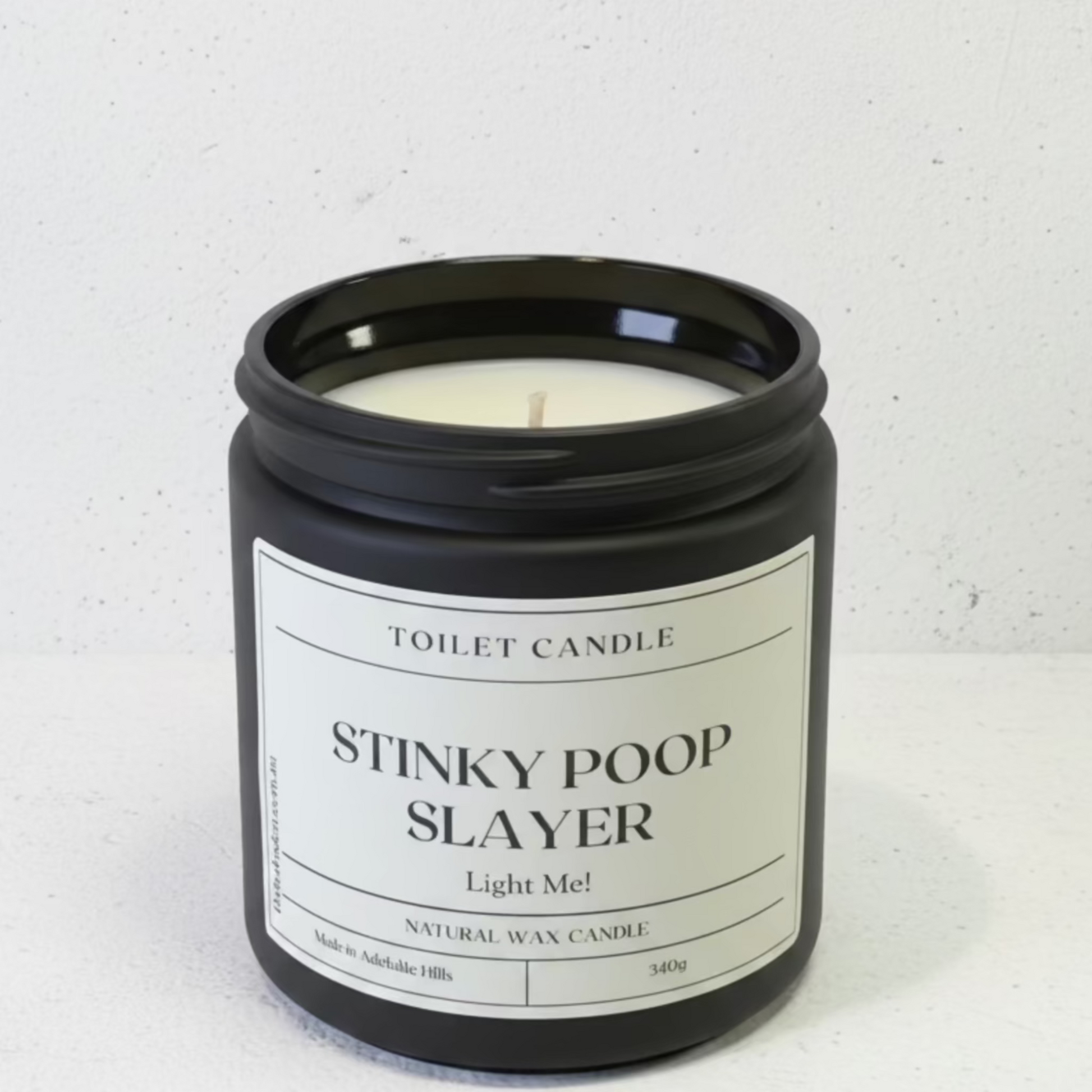 A black glass jar candle with a white label reading 'TOILET CANDLE STINKY POOP SLAYER Light Me!' on a light gray textured background.