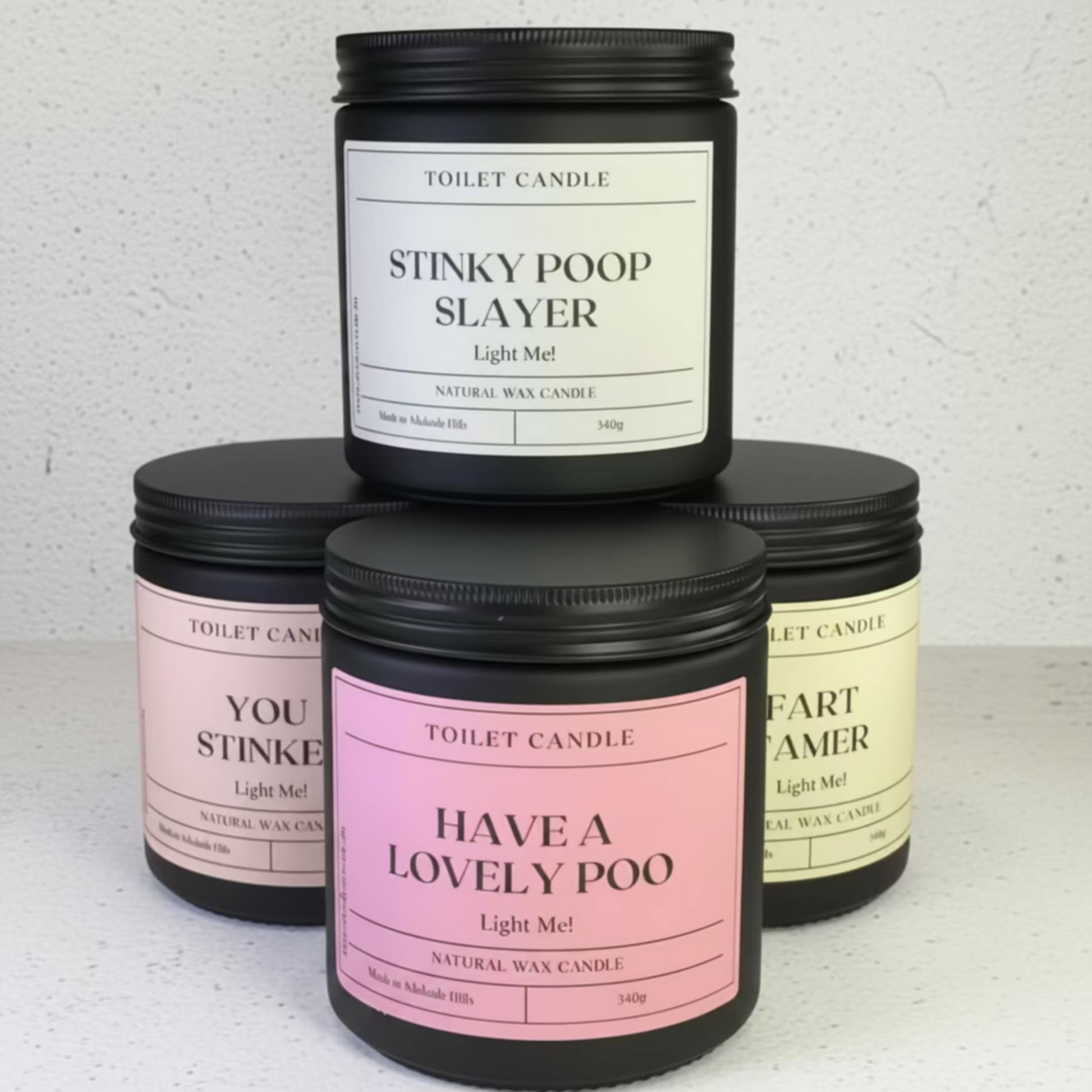 Four black glass  jar candles with black lids are stacked in a pyramid arrangement on a white speckled countertop against a white textured wall. The top candle has a white label reading 'STINKY POOP SLAYER' and the bottom center candle has a pink label reading 'HAVE A LOVELY POO'.